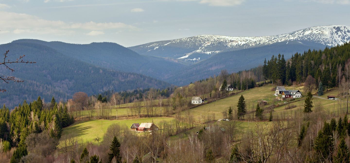 Krkonoše na jaře: louky a chalupy osady Levínek, za nimi zasněžený hřbet Kotle